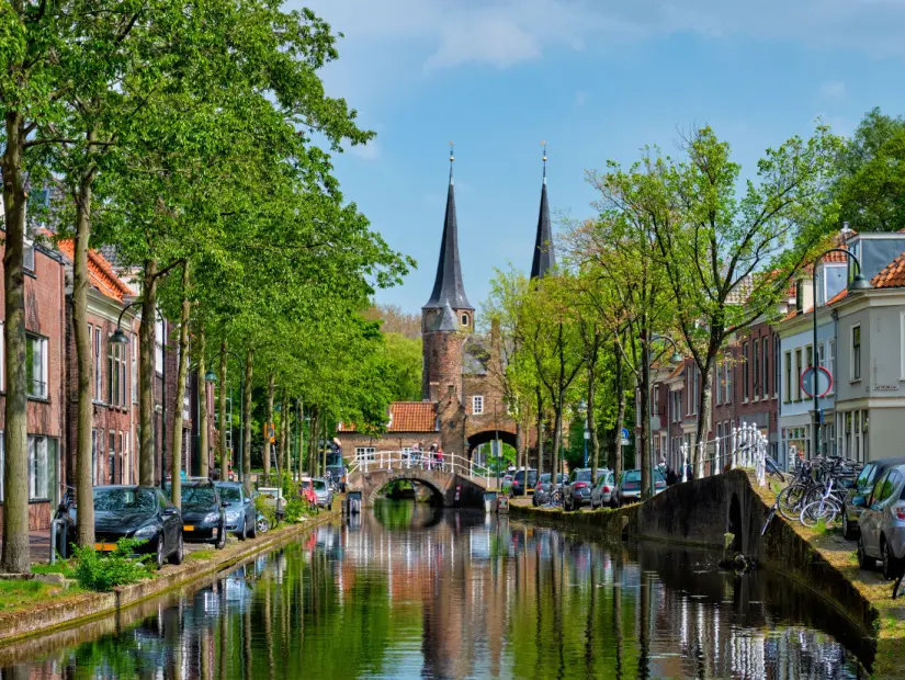 Doğu Kapısı Oostport ve birlikte park edilmiş araba ve bisikletlerin bulunduğu kanal ile pitoresk Delft şehir manzarası.